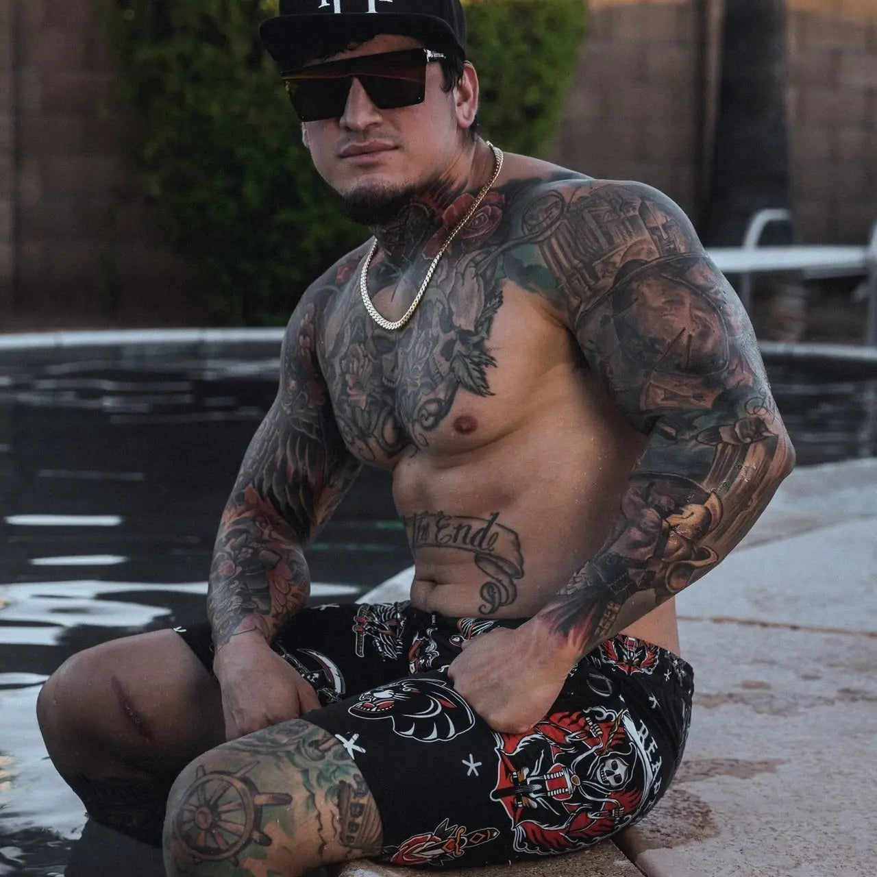 Tattooed man in swim trunks showcasing men’s boardshorts, motorcycle outerwear, and gloves