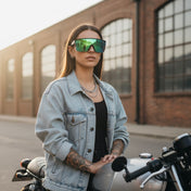 Woman wearing a denim jacket and reflective sunglasses standing next to a motorcycle.