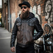 Man wearing a black leather jacket and cap standing in front of a graffiti-covered wall.