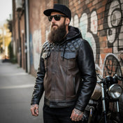 Man wearing a leather jacket and sunglasses standing next to a motorcycle with a graffiti-covered wall in the background.