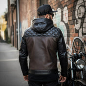 Person wearing a black leather jacket with quilted back standing next to a motorcycle against a graffiti-covered wall.