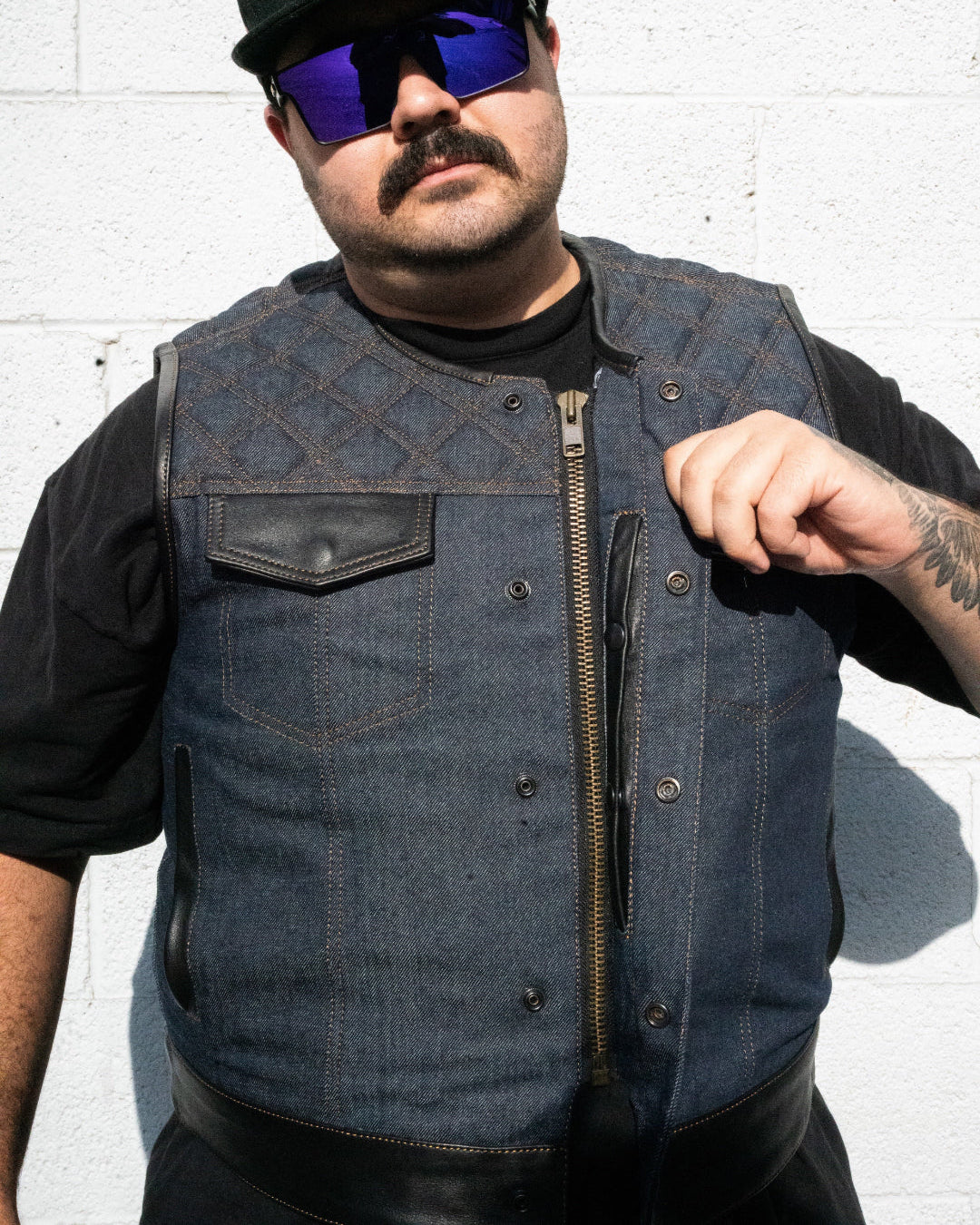 Man wearing a black cap, sunglasses, and a denim vest against a white brick wall.