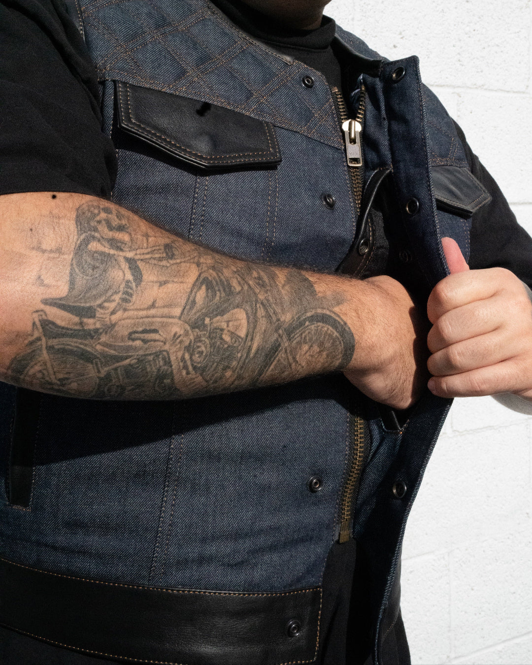 Man wearing a denim vest with tattoos on his arm, standing against a white brick wall.