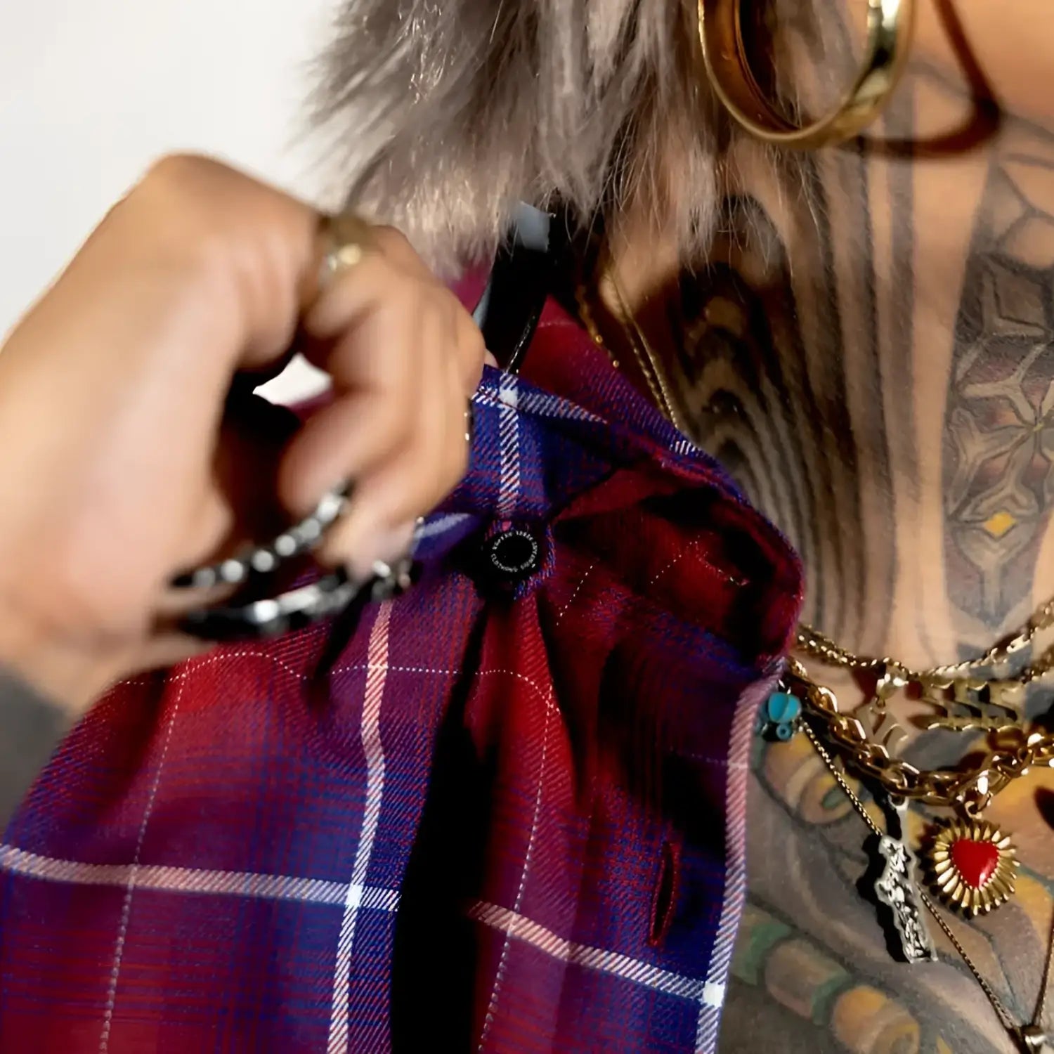 Person holding a red plaid bag with tattoos and jewelry on a neutral background