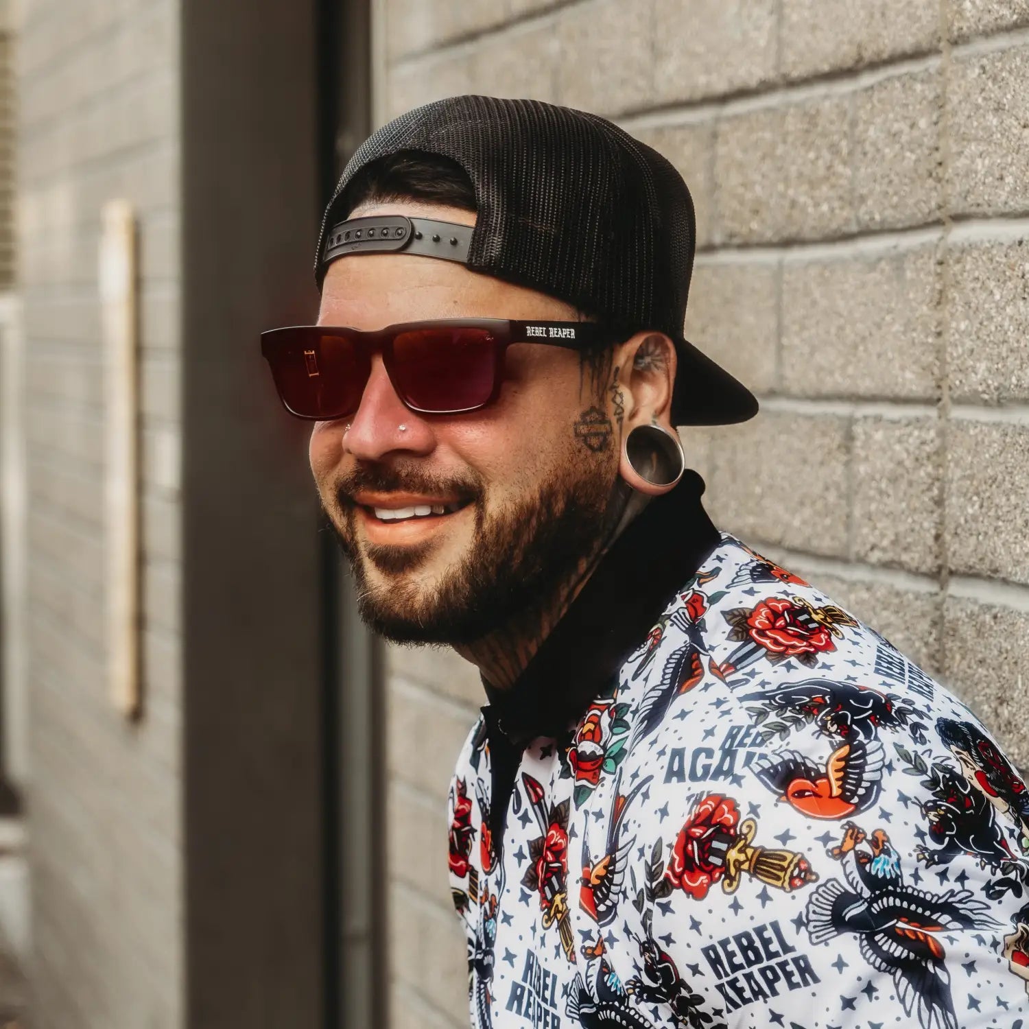 Smiling man with tattoos and sunglasses from Hustler Wayfarer Sunglass Collection featuring signature Rebel Reaper interchangeable arms