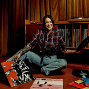 Person sitting on the floor surrounded by vinyl records in a room with wooden walls.