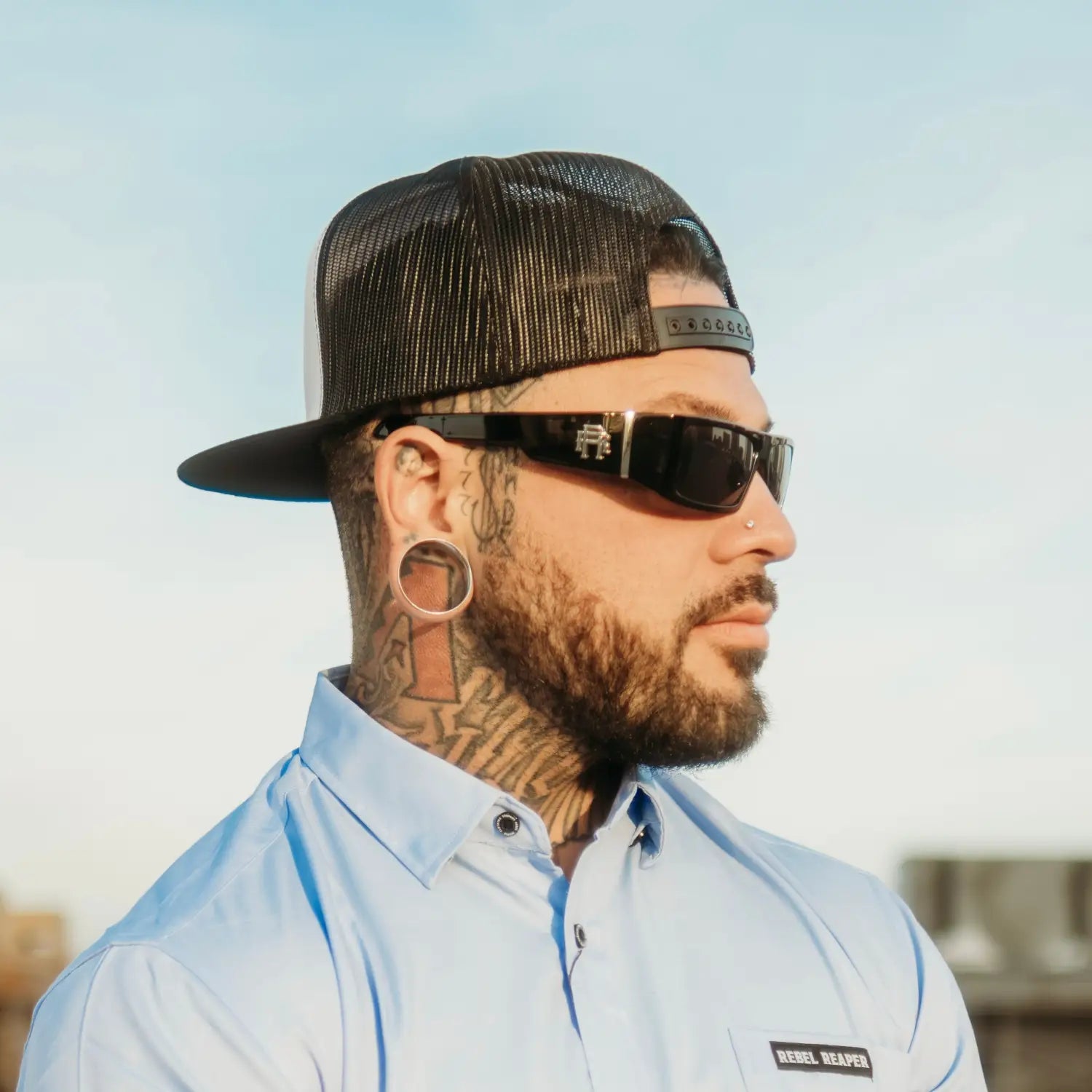 Man with tattoos, wearing sunglasses and a trucker hat from the Rebel Reaper Monogram Metal Rebel Reaper polarized sunglass collection